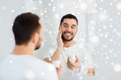 beauty, skin care, winter and people concept - smiling young man applying cream to face and looking to mirror at home bathroom over snow